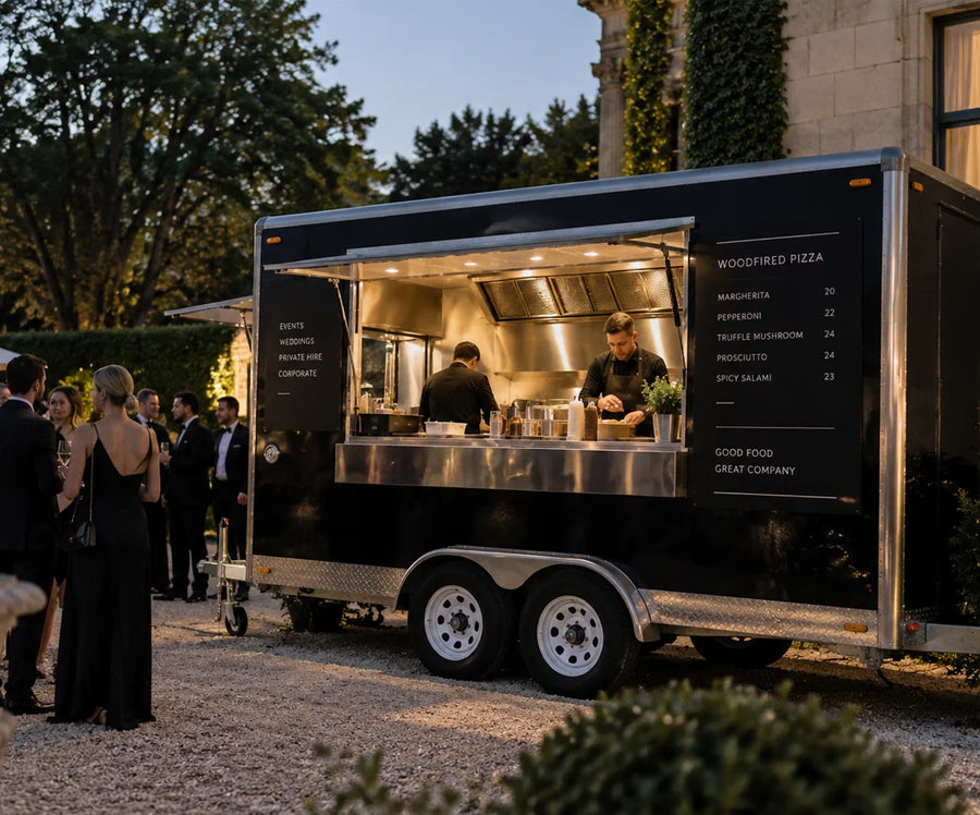Custom black tandem axle woodfired pizza trailer catering a luxury outdoor wedding with chefs preparing artisan pizzas